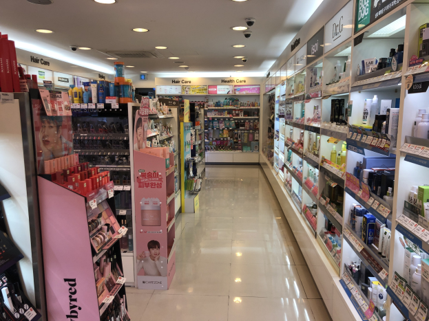 A cosmetics store with various hair care products displayed on racks, shelves, and the floor, featuring illuminated wall boards and overhead lighting.
