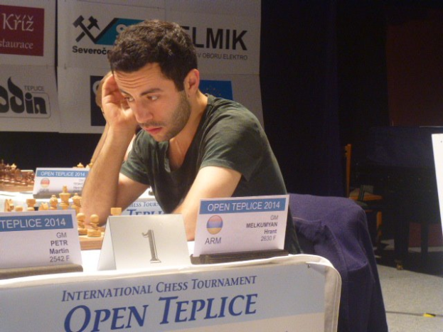 Ein Mann sitzt an einem Schachbrett mit aufgestellten Figuren, umgeben von Namensschildern und anderen Gegenständen, mit einem Banner im Hintergrund, auf dem "Open Chess Tournament 2014" steht.