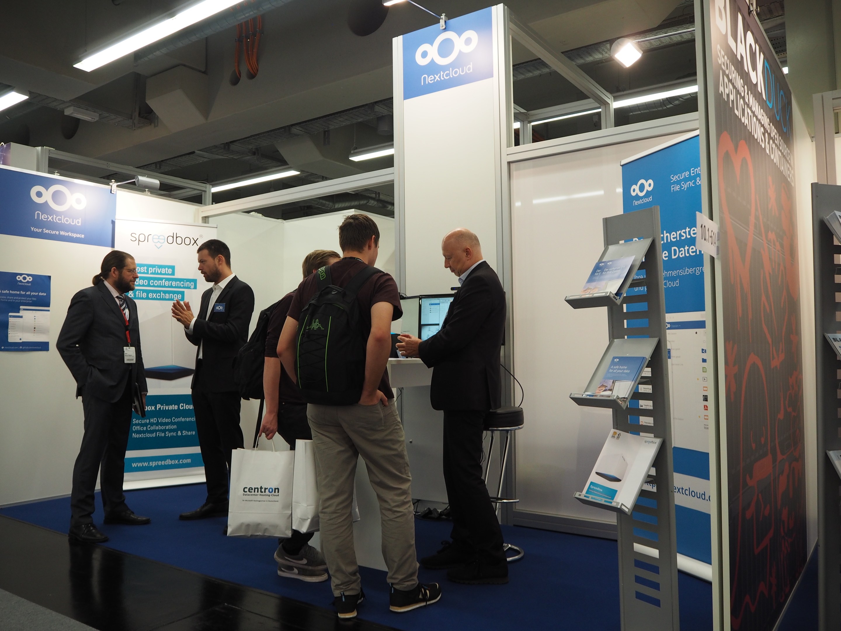 Menschen vor einem Messestand mit der Aufschrift "Ooo Nextcloud 2019", mit Bannern, einem Tisch mit einem Monitor, einem Stuhl, Deckenleuchten und Rohren.