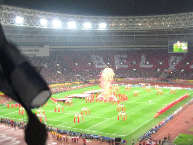 A stadium with floodlights, iron grills, spectators, standing individuals, advertisement boards, display screens, a security camera, flags, and decorative elements.