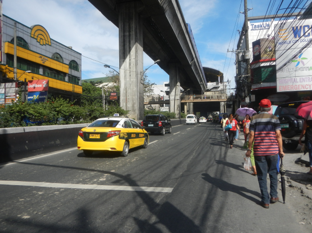 Ein gelbes Taxi fährt auf einer Stadtstraße mit hohen Gebäuden, Fußgängern auf dem Gehweg mit Regenschirmen, einer Brücke im Hintergrund und bewölktem Himmel mit Strommasten und Drähten.