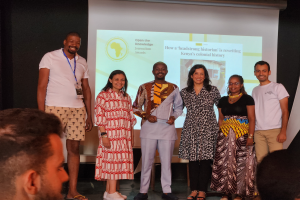 Eine Gruppe von Menschen auf einer B├╝hne, die fr├Âhlich in die Kamera l├Ącheln und posieren, mit einem Publikum davor und einem Bildschirm im Hintergrund, auf dem 'Open the Knowledge Journalism Awards 2019' angezeigt wird.