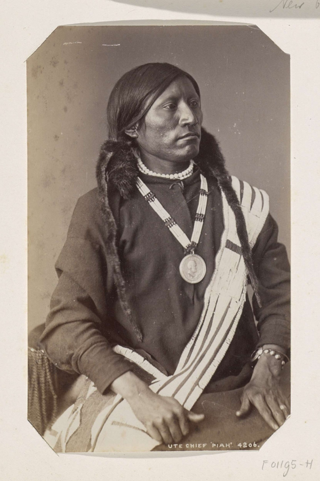 Schwarzes und weißes Foto eines amerikanischen Ureinwohner, identifiziert als Piah, Häuptling des Ute-Indianerstamms, der traditionelle Kleidung und Schmuck trägt, während er auf einem Stuhl sitzt.