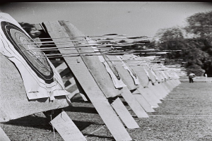 Schwarz-weisses Foto eines Bogenschiess-Ziels mit Pfeilen in der Mitte, umgeben von Bäumen und einer Person im Hintergrund, mit Boden sichtbar unten.