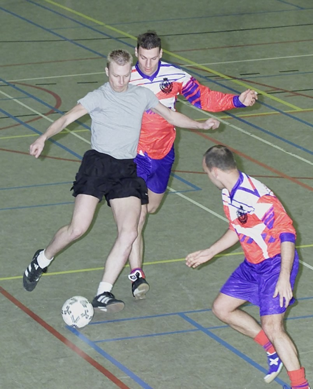 Drei Männer in Fußballtrikots und -schuhen spielen Futsal auf einem Court mit einem Ball im Spiel.