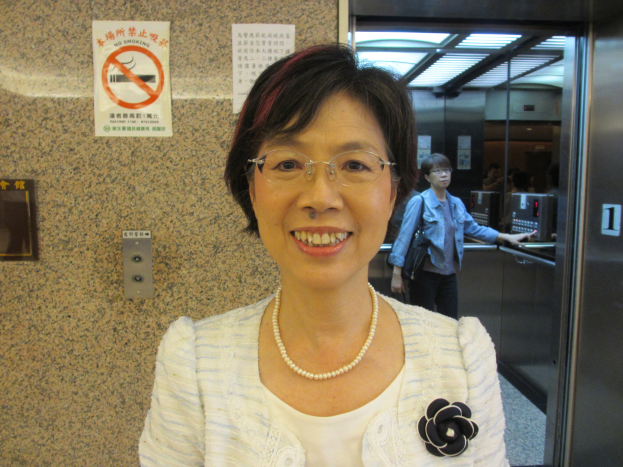 Eine Frau lächelt, während sie vor einem Aufzug steht, mit zwei Plakaten an einer nahen Wand und ein paar Menschen rechts von ihr.