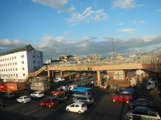 Vielbefahrene städtische Kreuzung mit Fahrzeugen, einer Brücke, Gebäuden und bewölktem Himmel mit Wasserzeichen.