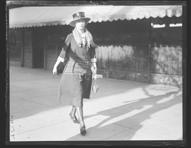 Eine Frau in einem Kleid und Hut geht die Straße entlang in Schwarz-Weiß, hält eine Handtasche und trägt hohe Absätze.