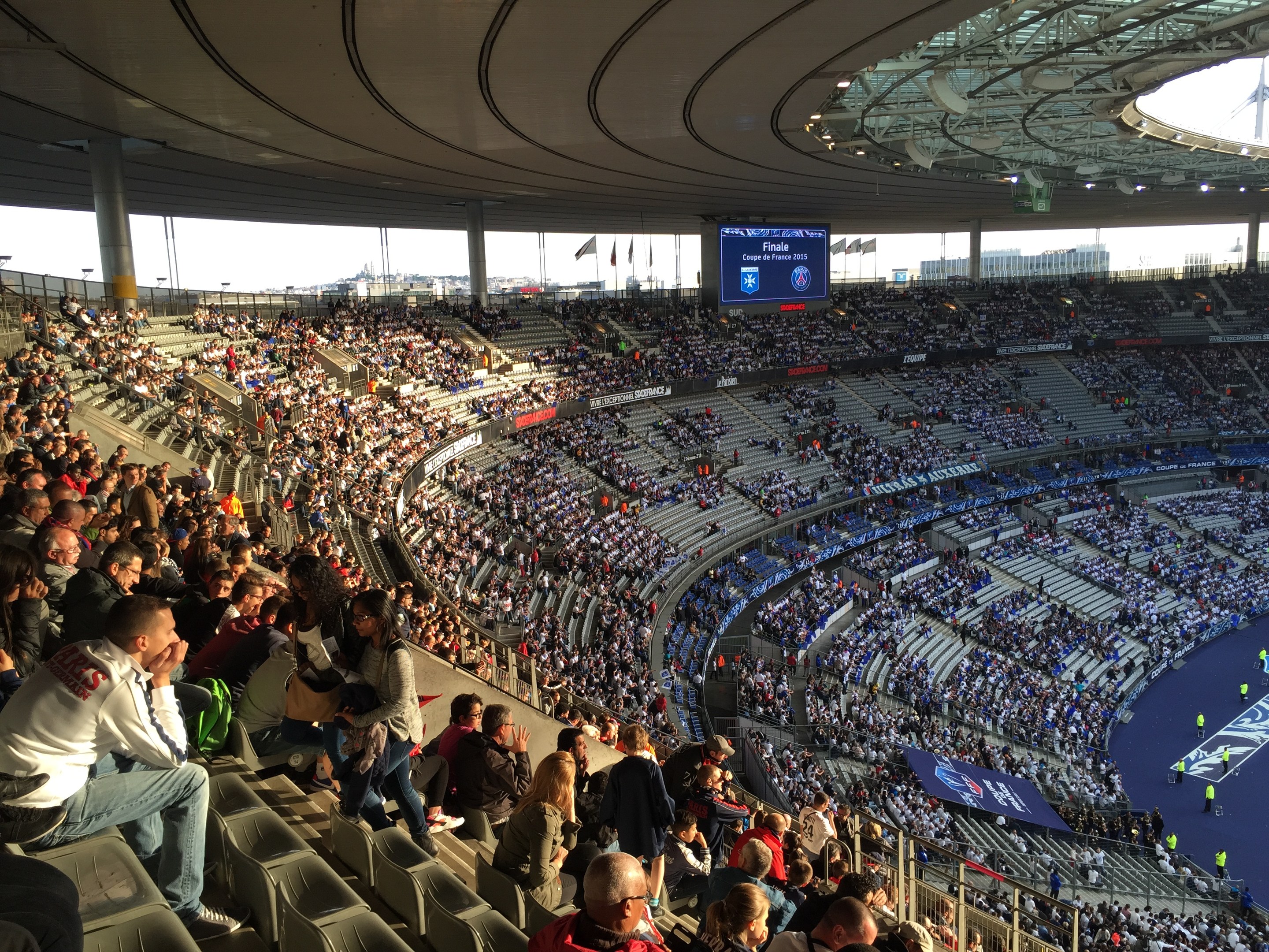 Große Menschenmenge in einem Stadion bei einem Fußballspiel mit einer Bühne, Fahnen, Stangen und einem Bildschirm im Hintergrund, identifiziert als Allianz Arena in München, Deutschland.