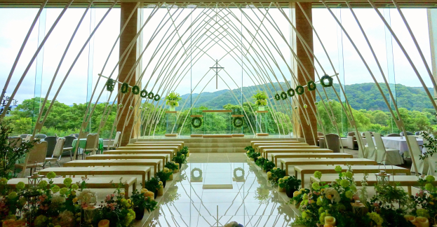 Eine Trauungszeremonie in einem großen Saal mit halbrunder Bestuhlung, Blumenschmuck und einem Hintergrund mit Bäumen, Hügeln und einem klaren Himmel.