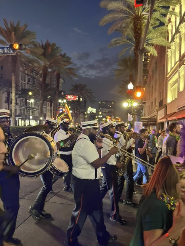 Eine Gruppe von Menschen in Kappen marschiert nachts eine Straße entlang und spielt Instrumente während eines Mardi Gras-Umzugs, mit Bäumen, beleuchteten Gebäuden und Texttafeln, die die Route säumen.