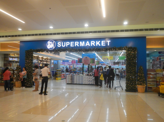 Gruppe von Menschen vor einem Supermarkt mit Einkaufstägern, einigen mit Einkaufstägern, mit einem Einkaufswagen, einem Topf mit einer Pflanze, Regalen, Schildern und einer Decke mit Leuchten.