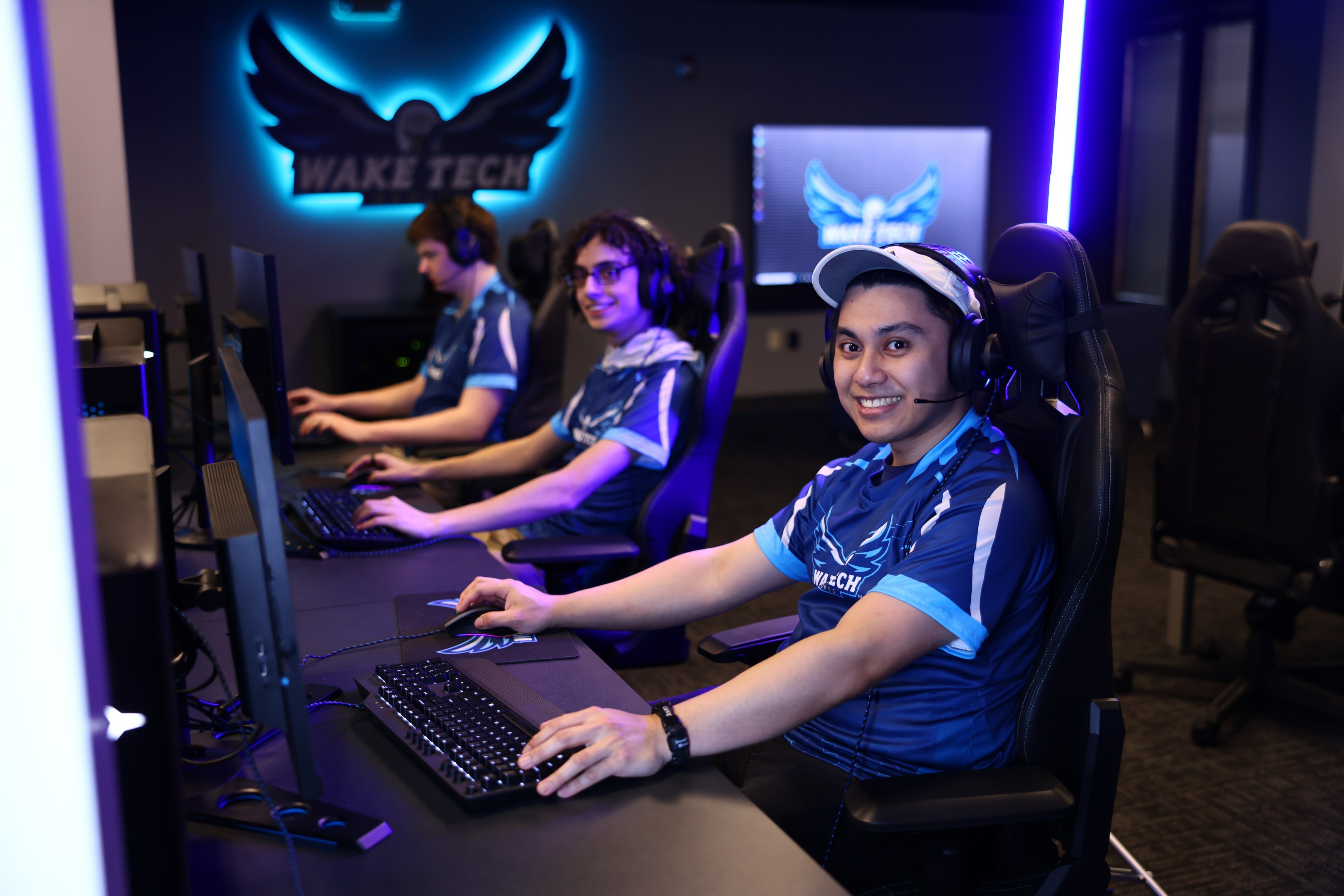 Eine Gruppe von Menschen in Headsets lächelt während sie bei einem Tech-Esports-Turnier arbeiten, mit Monitoren, Tastaturen und einer markierten Wand im Hintergrund.