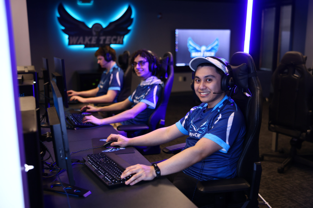 Eine Gruppe von Menschen in Headsets lächelt während sie bei einem Tech-Esports-Turnier arbeiten, mit Monitoren, Tastaturen und einer markierten Wand im Hintergrund.