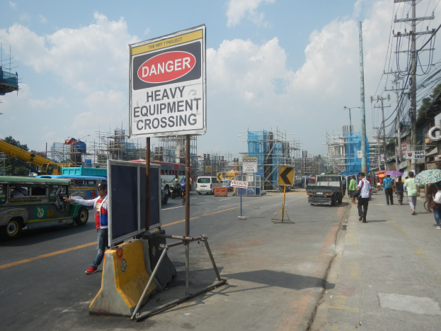 Busy street with a "Danger Heavy Equipment Crossing" sign, vehicles, pedestrians, electric poles, construction buildings, trees, and cloudy sky.