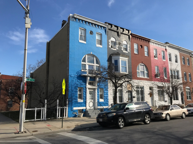 Eine Reihe bunter Häuser mit parkenden Autos auf einer Straße mit Bäumen, Pfählen, Brettern und einem Zaun unter einem bewölkten Himmel.