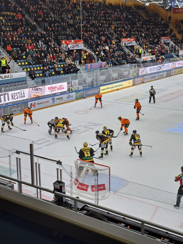 Eine Gruppe von Menschen, die Hockey auf einer Indoor-Eisbahn spielen, umgeben von einem Zaun, mit Zuschauern im Hintergrund und beleuchteten Bannern über ihnen.