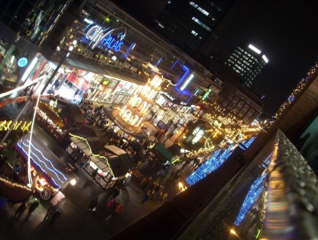Eine belebte nächtliche Stadtstraße voller Fußgänger, beleuchtet von Gebäuden und Marktständen gegen einen dunklen Himmel.
