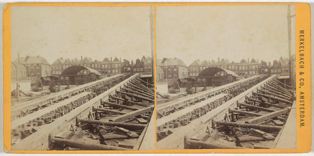 Ein altes Schwarz-Weiß-Foto einer Bahnschiene mit Gebäuden im Hintergrund, Text auf der rechten Seite, der "Merkelbach & Co. Amsterdam" lautet, und verstreuten hölzernen Gegenständen im Vordergrund.