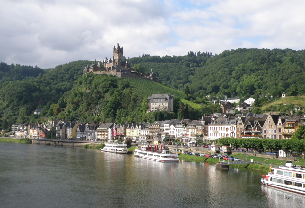Ein malerischer Blick auf den Rhein in Deutschland mit einer Burg auf einem Hügel im Hintergrund, Booten auf dem Fluss und Fahrzeugen auf einer näheren Straße sowie einem bewölktem Himmel.
