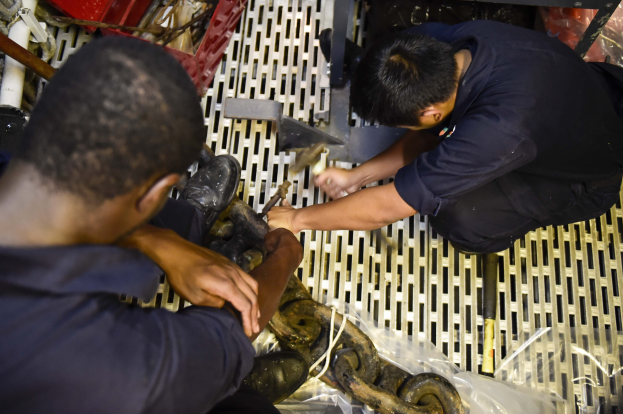 Zwei Männer in einer Fabrik, die an einer Maschine arbeiten und Werkzeuge sowie Rohre und andere Gegenstände um sie herum halten.