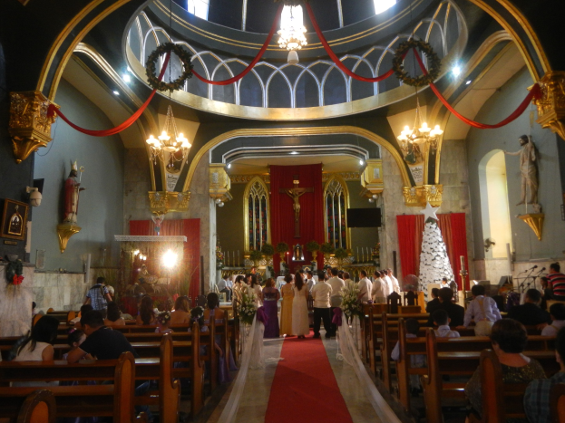 Eine Trauungszeremonie in einer Kirche mit einem roten Teppichgang, Menschen auf Bänken sitzend, einige stehend mit Blumensträußen, Wände mit Statuen und Lichtern geschmückt und eine dekorierte Decke mit Kronleuchtern.