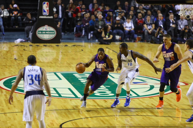 Basketball-Spiel zwischen den Phoenix Suns und Dallas Mavericks mit Spielern auf dem Feld und Zuschauern in den Rängen, Basketballkorb sichtbar oben rechts.