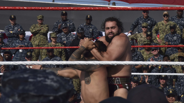 Zwei Wrestler in einem Ring, umgeben von Seilen, die sich gegenüberstehen, mit einer Zuschauermenge und einer roten Wand im Hintergrund und offener Himmel.