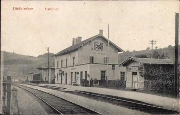 Black and white photograph of Dielkirchen train station in Germany with a train on the tracks, surrounded by houses, utility poles, trees, and people, with hills and a clear sky in the background.