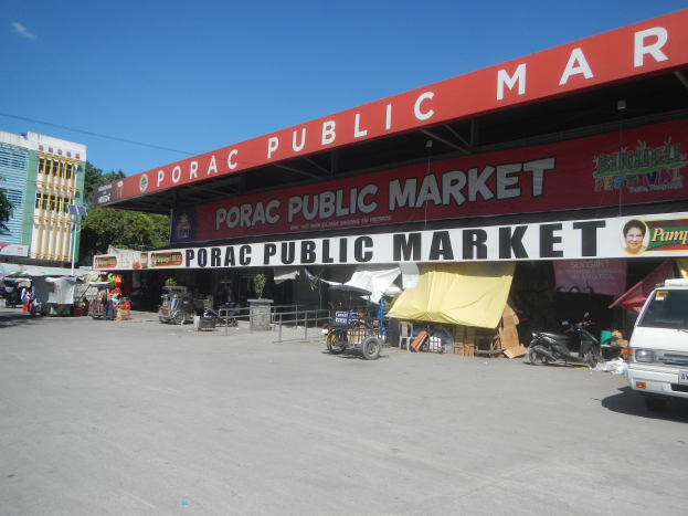 Eine belebte Straßen Szene auf dem Porac Public Market in Manila, Philippinen, mit Fahrzeugen, Marktständen, Gebäuden, Bäumen und einem klaren blauen Himmel.