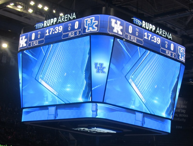 Großer Bildschirm auf einem Basketballfeld, der "Kentucky Wildcats vs Florida State Seminoles" anzeigt, mit Zuschauern auf den Tribünen und Beleuchtung von oben.