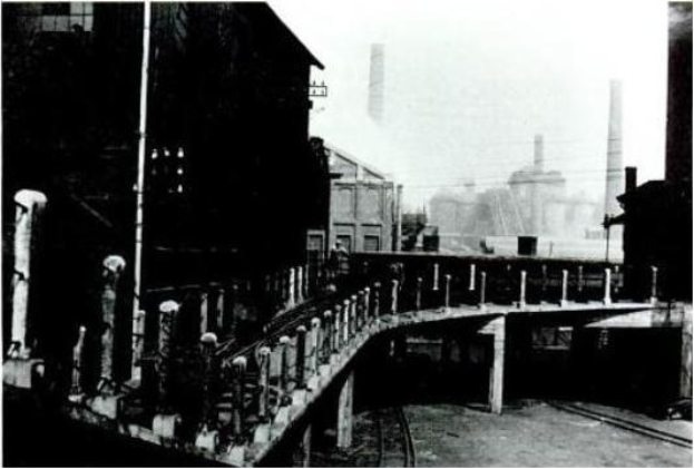 Black and white photo of a bridge over train tracks, with buildings on either side and a factory in the background, featuring railings and poles along the sides.