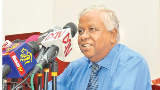 Ein Mann in einem blauen Hemd und Krawatte sitzt an einem Tisch mit Mikrofonen und lächelt, während er eine Erklärung abgibt.