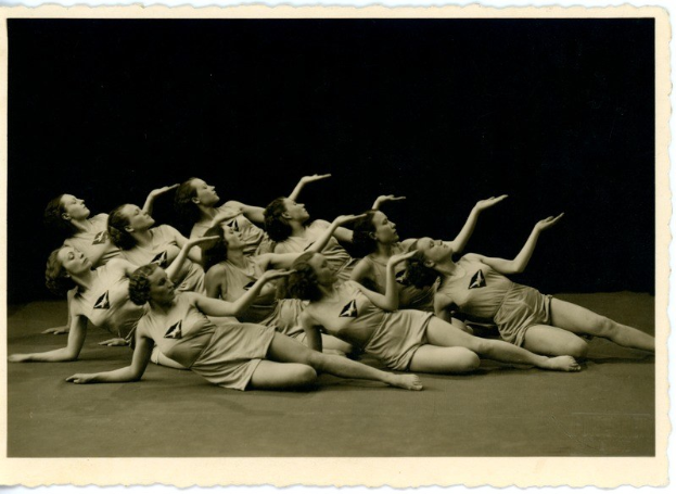 Schwarz-weiß-Foto einer Gruppe von Frauen, die synchronisierte Boden-Gymnastik in passenden Outfits durchführen, mit einem Strahlen in die Kamera blicken und verschiedene Posen einnehmen, gegen einen unscharfen, verträumten Hintergrund.