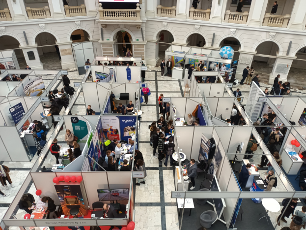 Eine grosse Gruppe von Menschen auf einer internationalen Handelsmesse mit Ständen, Bannern, Tischen, Stühlen, Ballons und einem Gebäude mit Bögen, Säulen, Geländern und einem Bildschirm im Hintergrund.