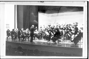 Ein Schwarz-Weiß-Foto eines Dirigenten in Anzug und Krawatte, der auf einer Bühne vor einem Orchester steht, das verschiedene Instrumente spielt, während das Publikum aufmerksam zuhört.