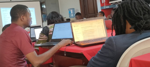 Group of people seated around a table with laptops during an internet usage training session, with a screen, door, and water can visible in the background.