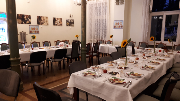 Ein Speisesaal mit mehreren Tischen und Stühlen, dekoriert mit Blumenvasen, Besteck, Lebensmitteln und Fotorahmen an der Wand, mit Vorhängen, Fenstern, einem Kronleuchter und einem Getränkeautomaten auf der linken Seite.