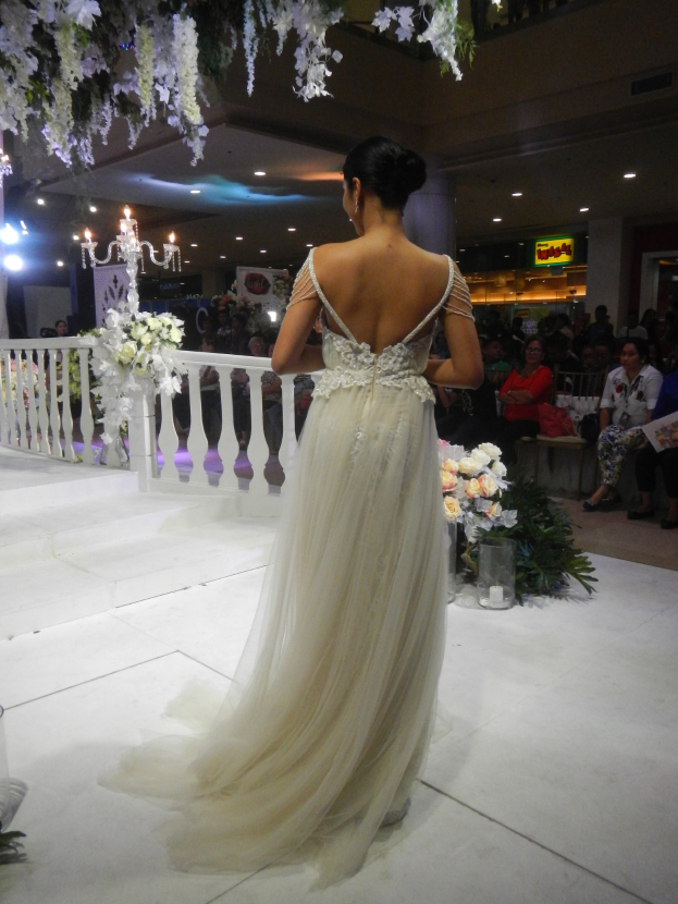 Eine Frau in einem Hochzeitskleid läuft über einen Laufsteg, umgeben von sitzenden Zuschauern, einer Reling, einem Kronleuchter, Blumen, Blättern und anderen Gegenständen, unter einer Decke mit Lichtern und Blumen.