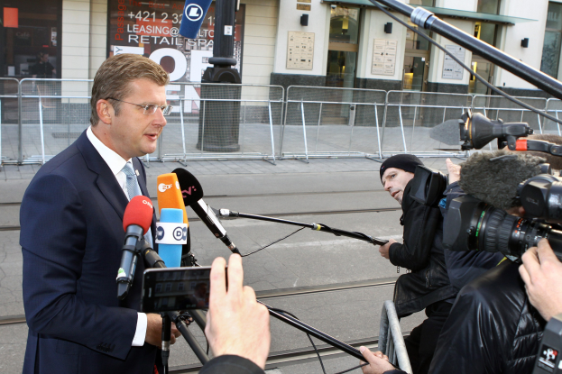 Ein Mann in Anzug steht vor Reportern mit Kameras und Mikrofonen, mit einem Metallzaun, einem Pfahl, einem Müllcontainer, Brettern mit Text, einem Gebäude mit Fenstern und Papieren an einer Wand.