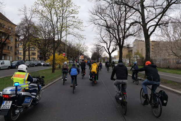 Eine Gruppe von Menschen, die auf Fahrrädern eine von Bäumen gesäumte Straße entlangfahren, begleitet von einem Polizeibeamten auf einem Motorrad.