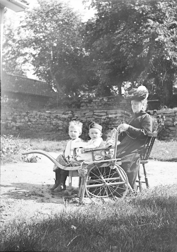 Schwarz-weiß-Foto einer älteren Frau in einem Kleid und Hut, die in einem Rollstuhl sitzt und von zwei Kindern umgeben ist, draußen vor einer Wand, Bäumen und Himmel