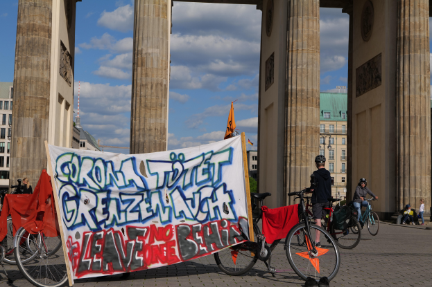 Eine Gruppe von Menschen auf Fahrrädern, die Helme tragen, fährt vor dem Brandenburger Tor in Berlin, Deutschland, mit einer Fahne und Gebäuden im Hintergrund bei bewölktem Himmel.