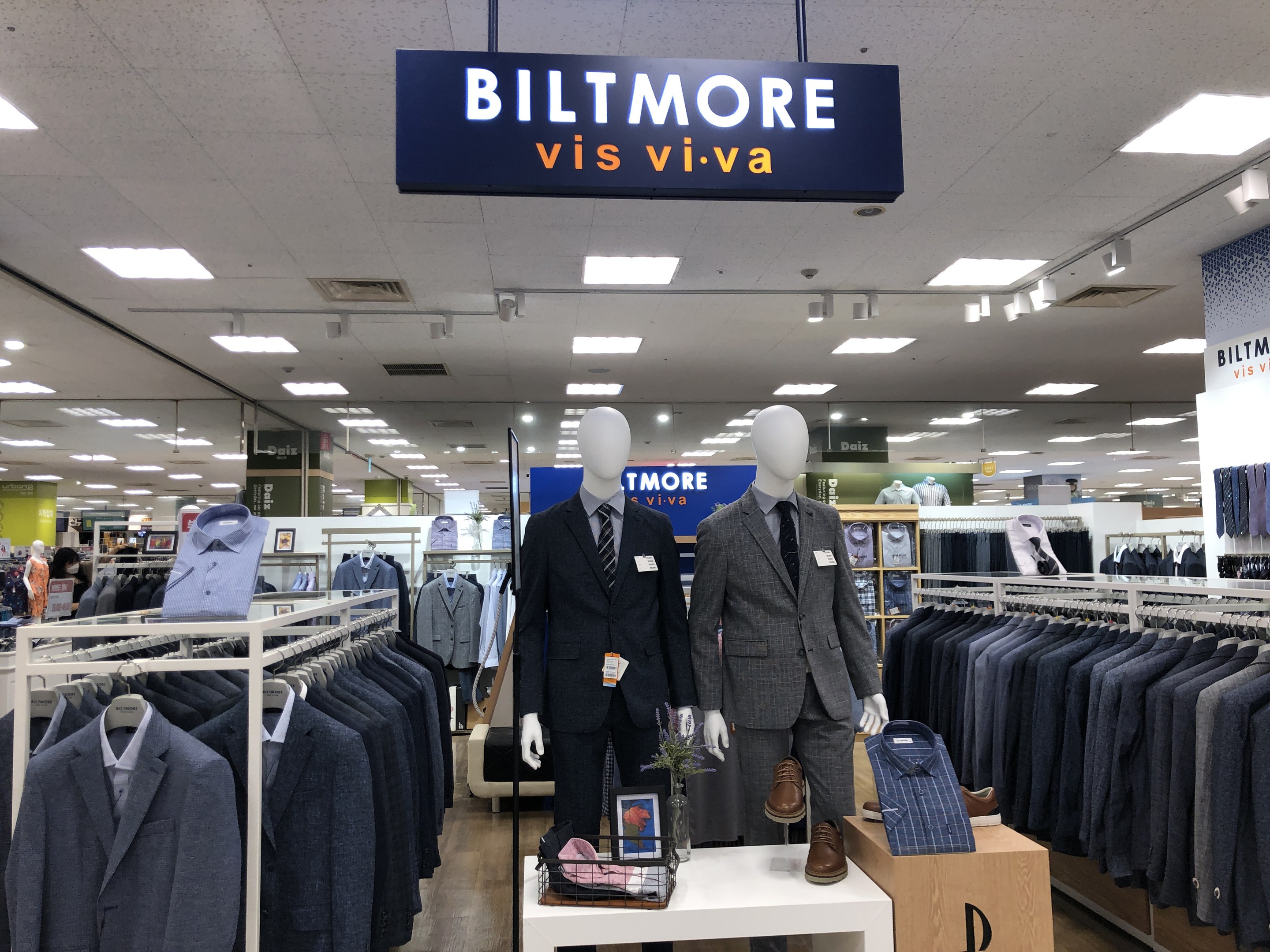 Innenansicht eines Biltmore Vis Viva Bekleidungsgeschäfts mit Anzügen auf Schaufensterpuppen, aufgehängten Kleidungsstücken, einem Tisch mit einer Blumenvase und einer Texttafel an der Decke.