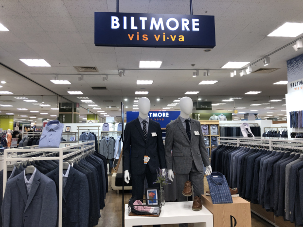 Innenansicht eines Biltmore Vis Viva Bekleidungsgeschäfts mit Anzügen auf Schaufensterpuppen, aufgehängten Kleidungsstücken, einem Tisch mit einer Blumenvase und einer Texttafel an der Decke.