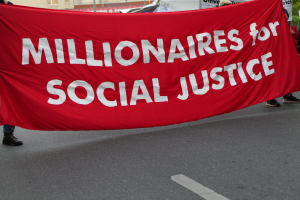 Group of people walking down a street holding a red banner reading "Millionaires for Social Justice" with buildings, poles, trees, and a clear blue sky in the background.