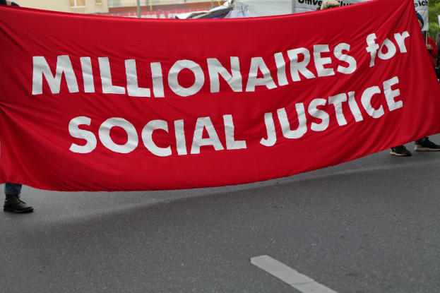 Group of people walking down a street holding a red banner reading "Millionaires for Social Justice" with buildings, poles, trees, and a clear blue sky in the background.