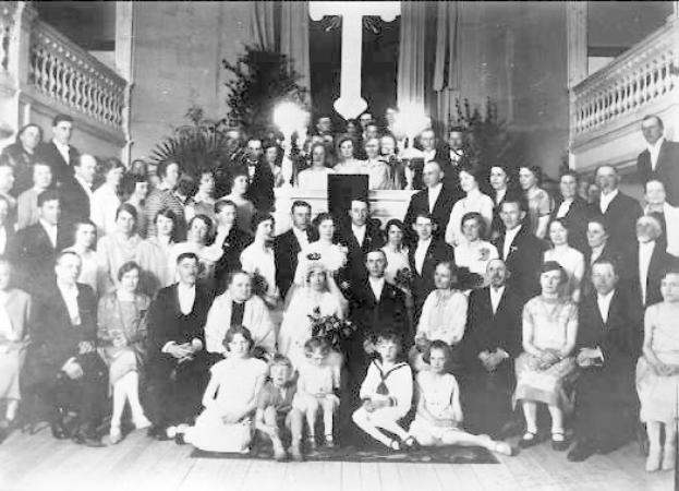 Schwarz-weißes Foto einer großen Hochzeitsgesellschaft aus den 1930er Jahren, die in einem Raum mit Pflanzen, Geländern und einer Wand im Hintergrund posiert.