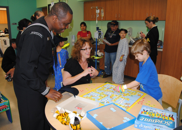 Gruppe von Menschen, die ein Brettspiel an einem Tisch mit Spielzeugen und Schachteln in einem Raum mit Schränken, Salzflaschen auf einem Ständer, einer Tür und einer Wand spielen.