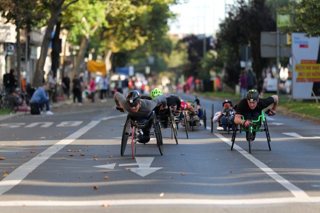 Eine Gruppe von Menschen in Rollstühlen, die eine Straße entlangrasen, Helme tragen und von Bäumen, Gebäuden und anderen Menschen umgeben sind, mit einem unscharfen Hintergrund, der Bewegung andeutet.